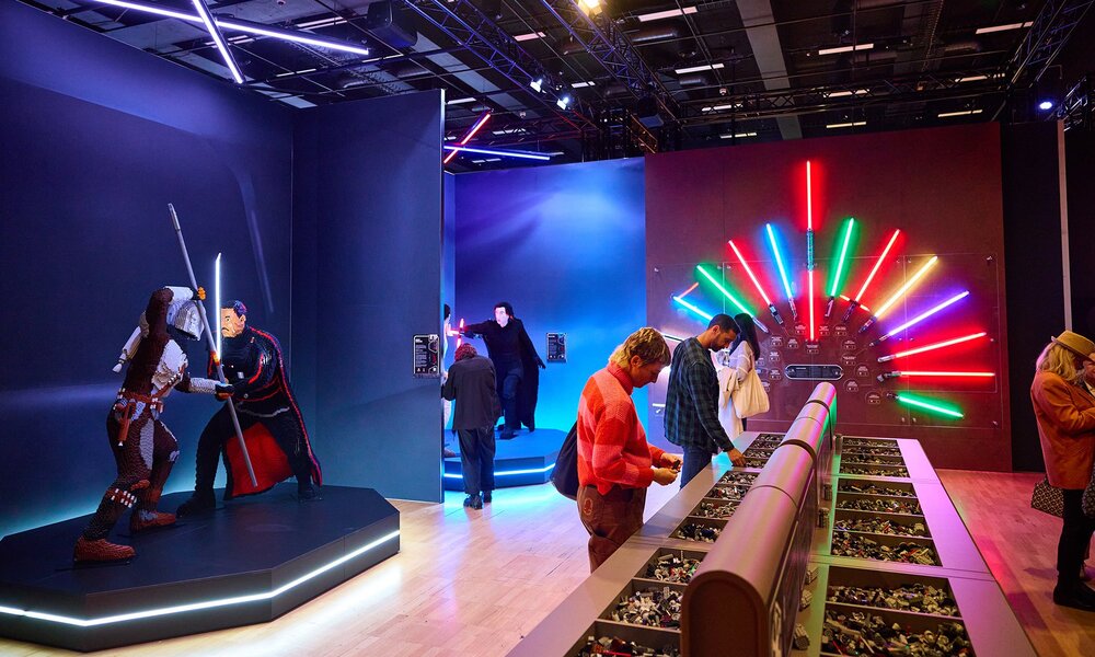 A Star Wars LEGO exhibition with characters fighting with lightsabers and people looking through tubs of bricks.