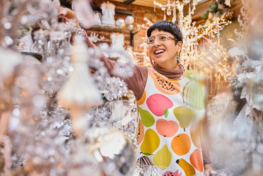 A market stallholder wearing a colourful apron decorated with pictures of fruit, surrounded by silver tinsel Christmas decorations.