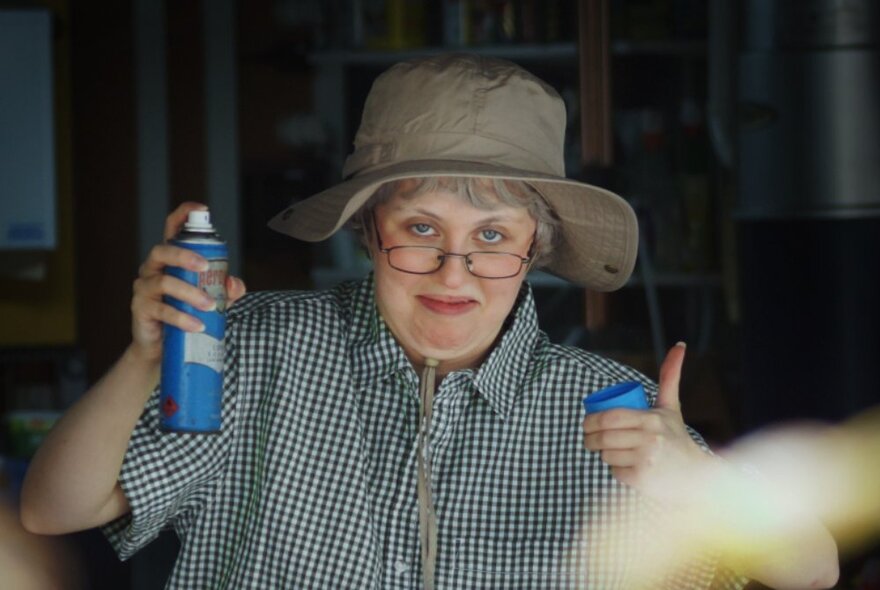 Performer wearing an old hat, glasses and shirt, holding a can of flyspreay and giving a thumbs up.