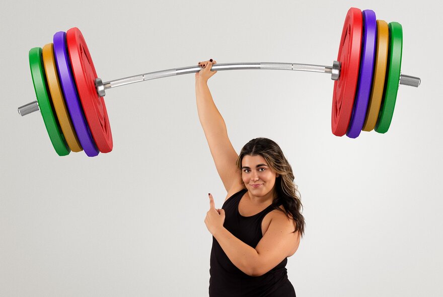 Comedian Ainslie with a large set of coloured novelty dumbells, lifted with one arm, above her head with her other hand pointing up looking smugly. 