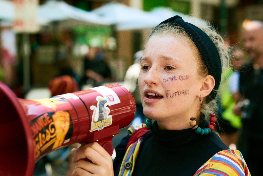 A woman protester with writing on her cheek and colourful clothes on, talking into a megaphone covered in stickers.