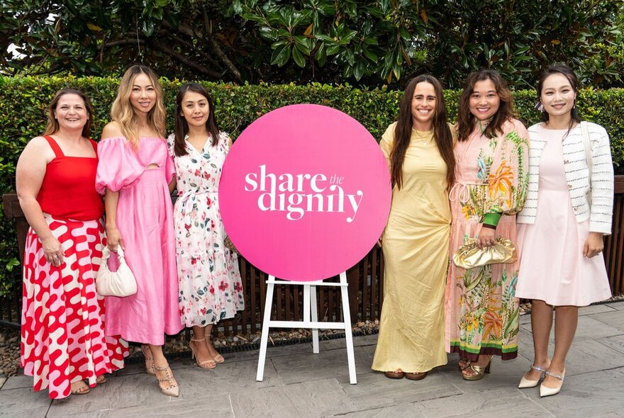 A pink 'Share the Dignity' sign with three women standing either side in dresses.