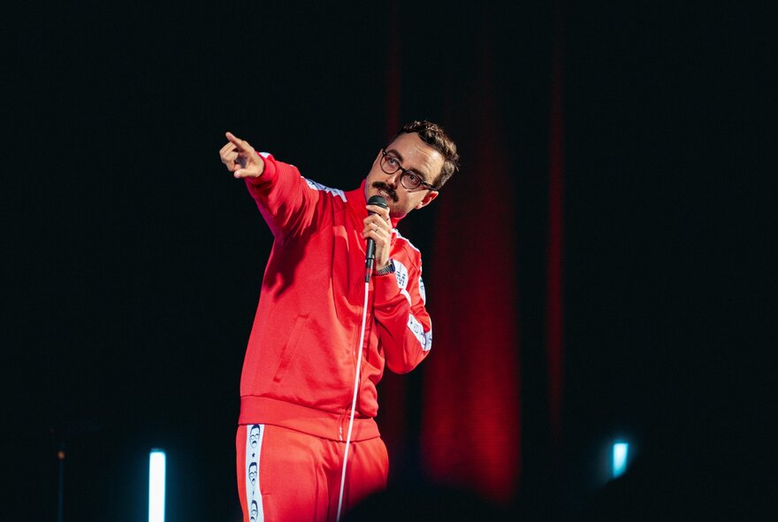 A comedian on stage, wearing a bright red track suit and glasses, pointing.