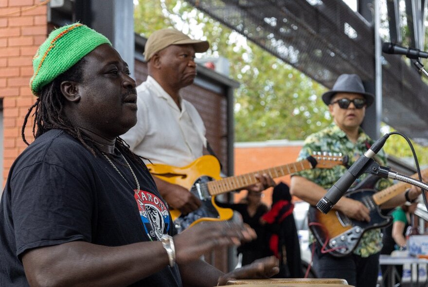 Three musicians of different nationalities wearing hats and playing their instruments on an outdoor setting. 