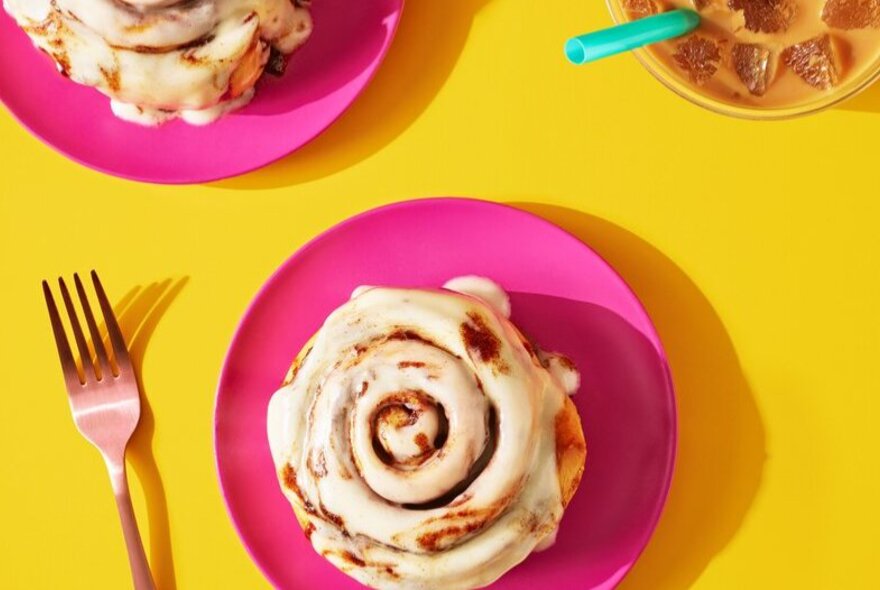 Overhead view of a cinnamon bun with frosting on a pink plate, set on a yellow table top, a fork alongside.
