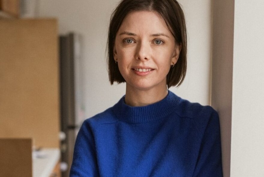 Young woman with brown bob hairstyle, wearing a cobalt blue turtleneck jumper and leaning against a wall, interior.