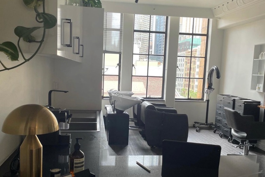 Interior of hair salon with club chairs, hair-washing basins, tables and tall windows on the far wall.