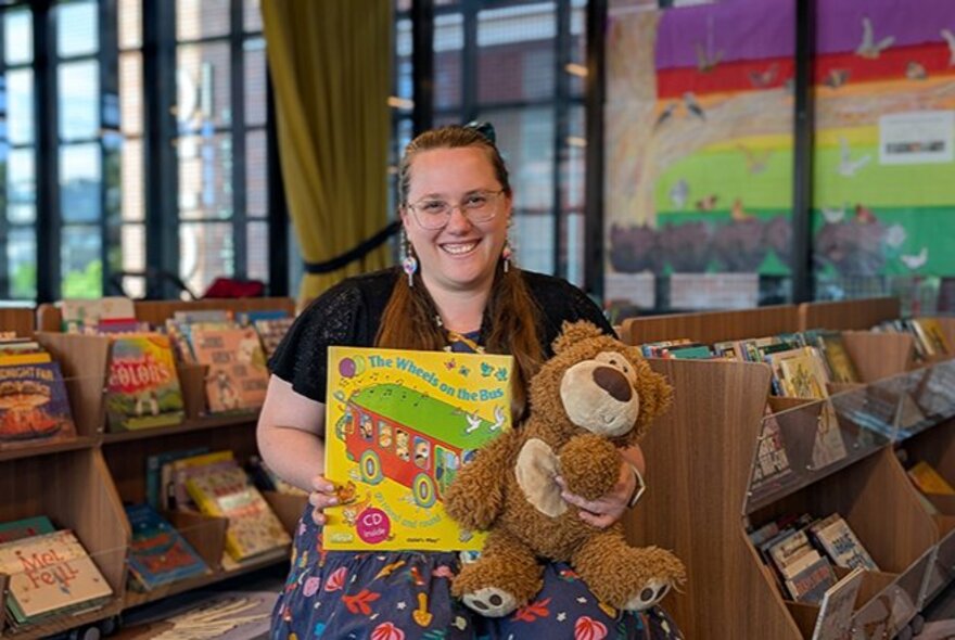 A smiling person seated in front of the children's section of a library, holding up a picture book and teddy bear in her lap.