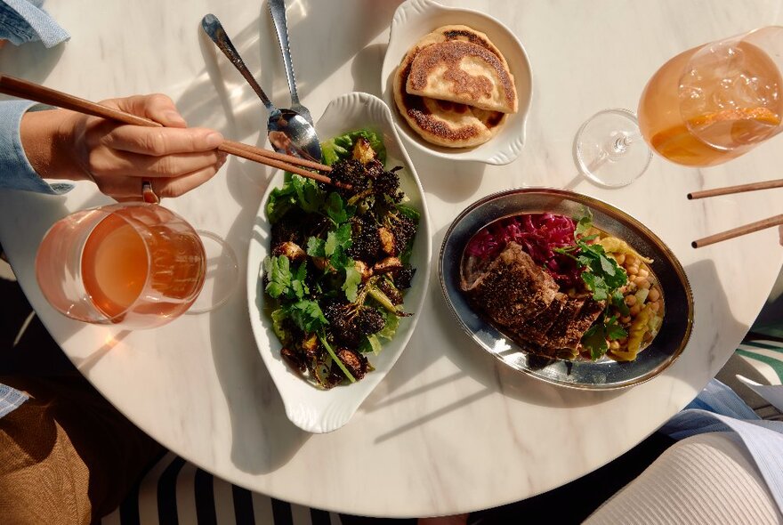 Looking down on a selection of Asian inspired dishes, and a side dish of roti, plus drinks, on a marble table top; hands leaning in with chopsticks.