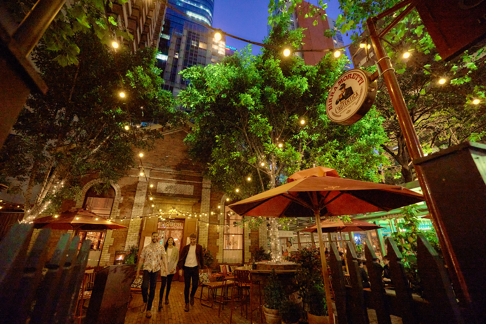 Three friends walking through a courtyard with trees, fairy lights and umbrellas outside a brick restaurant.