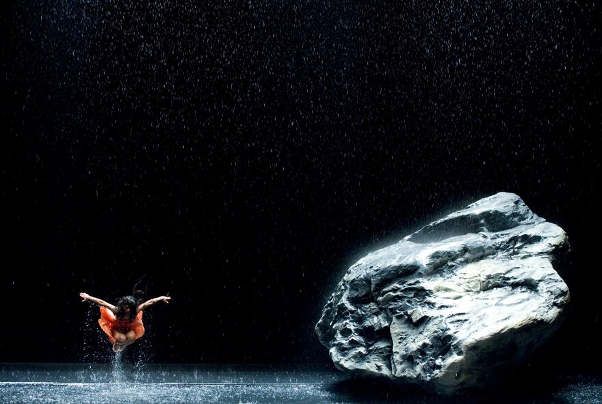 A dancer in an orange costume leaps in a shallow pool of water next to a large gray rock in a dark, rain-like setting. A still from the documentary film 'Pina'.