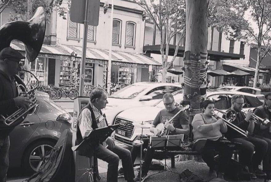 A black and white photo of a group of musicians playing music on the roadside, cars parked on the street behind.