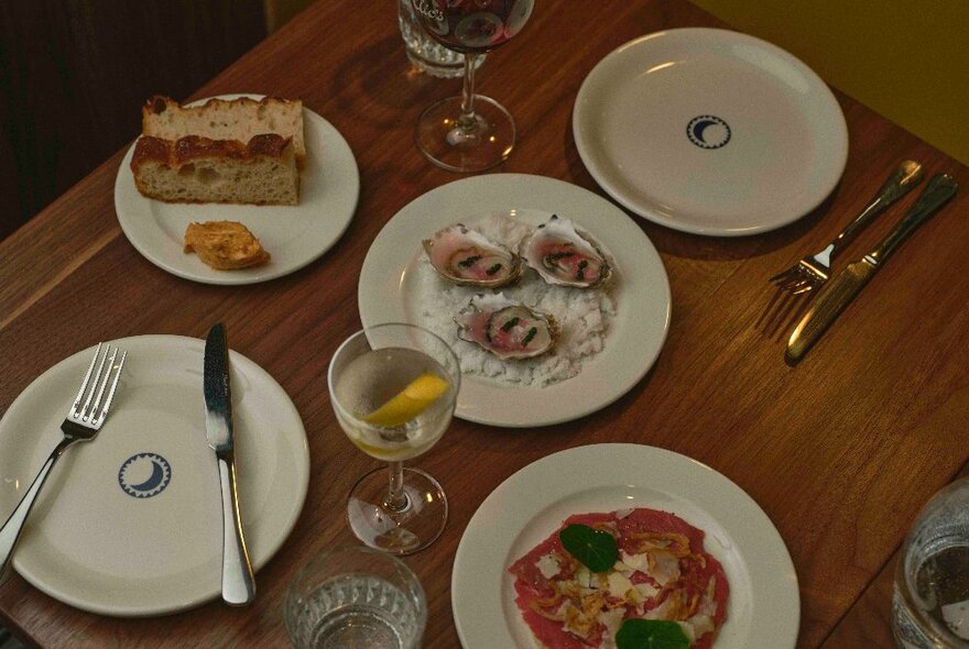 A dimly lit selection of Italian dishes, arranged on a wooden table with empty plates, wine glasses and cutlery.