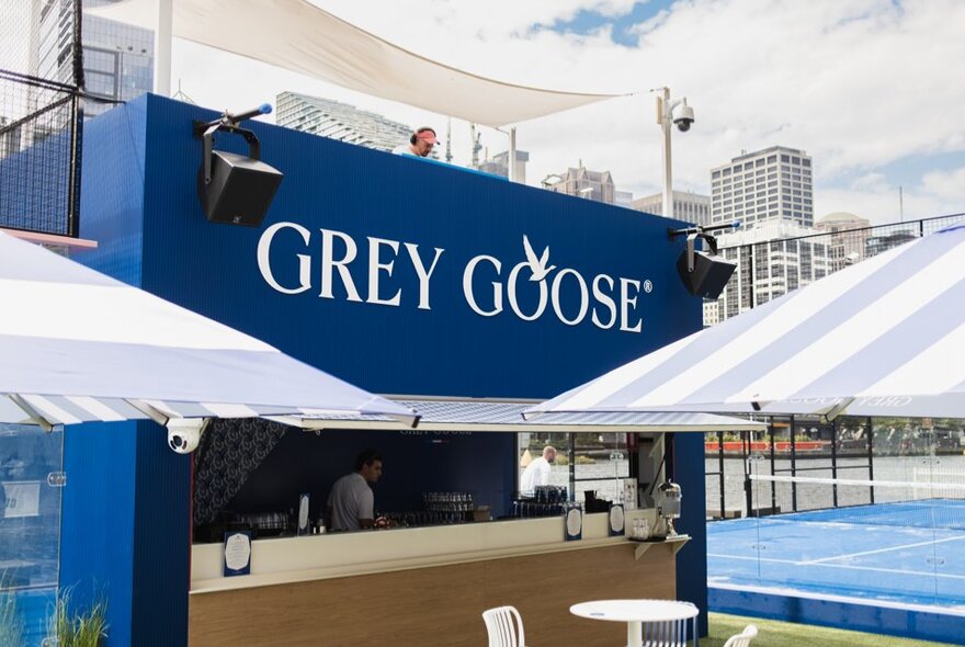 An open air bar with blue 'Grey Goose' signage and pale blue and white umbrellas in front of it; a blue padel court behind.