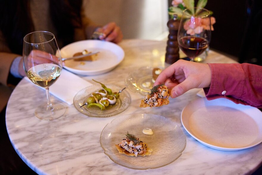 Someone lifting a cracker off a plate in a restaurant, with glasses of wine and other tapas dishes on the table.