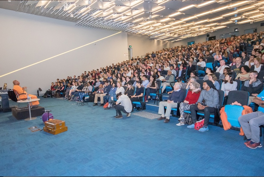 An auditorium with bright blue carpet, every seat filled with happiness workshop participants.