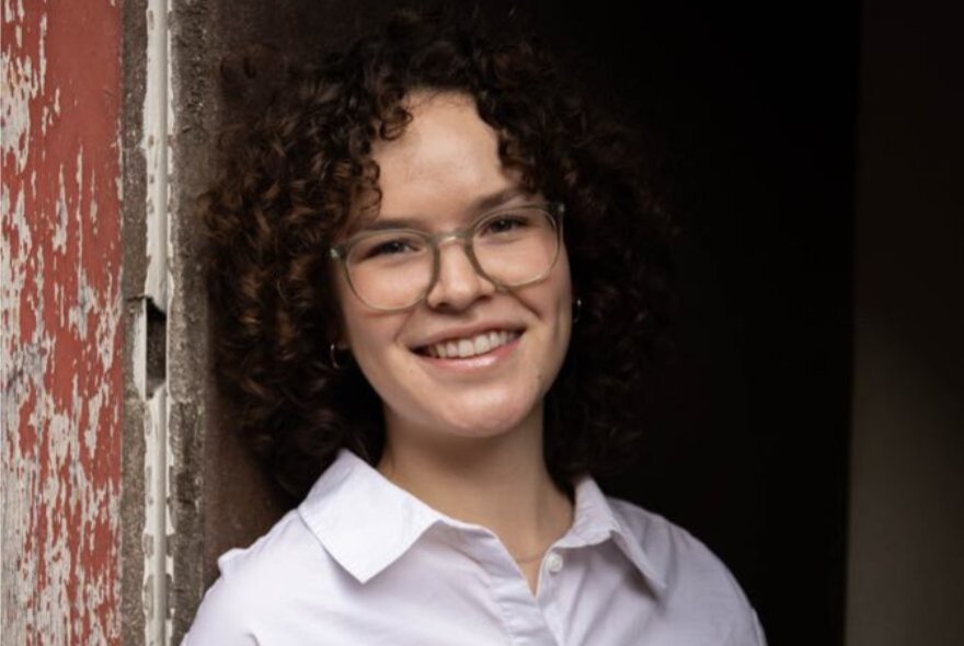Young writer Bianca Lucchesi, leaning against a doorway, smiling, wearing glasses and a white shirt.