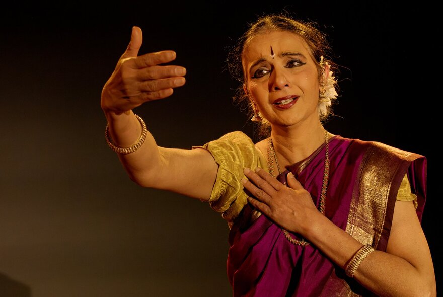 An Indian woman wearing a magenta sari with gold trim, traditional jewellery and face adornments, on stage.