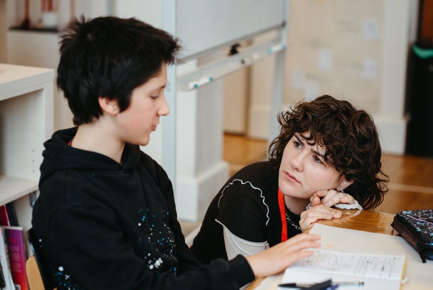 A teachers bobs down next to a seated student and listens intently. 