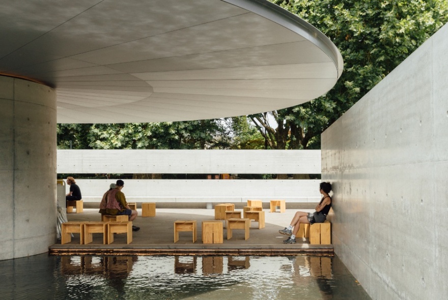 Inside MPavilion under the umbrella canopy, people sitting on small wooden stools. 