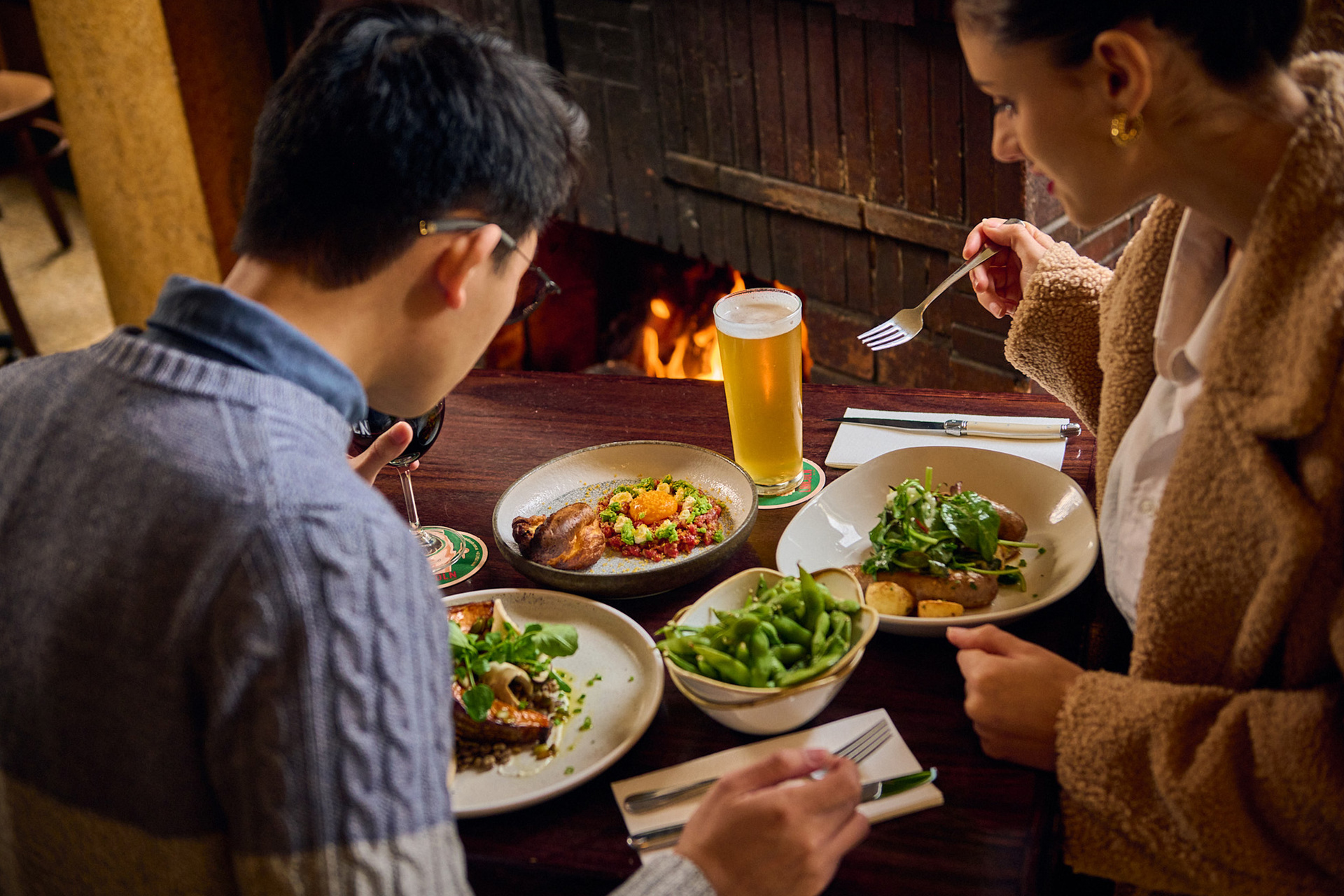  a couple are seated next to a fireplace in a pub they are eating edamame beans and steak and have a glass of beer on the table