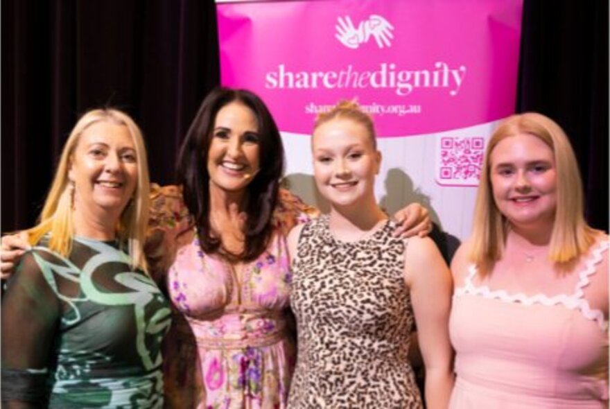 Four smiling women standing together in front of a pink 'share the dignity' sign.