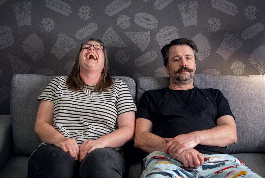 A man and a woman sit next to each other on a grey couch, the man with a dissatisfied look on his face, the woman with her head back laughing.