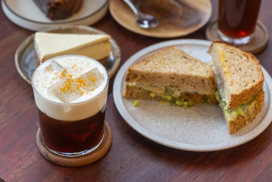 Cream and orange zest topped iced coffee on a wooden table with sandwich and cake.