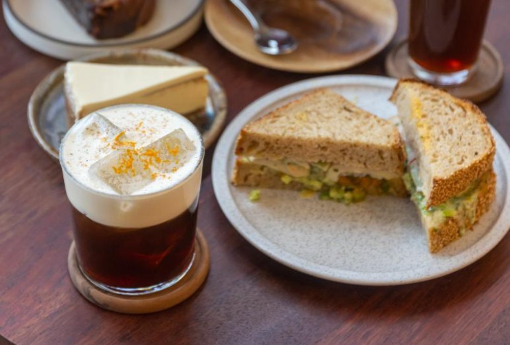 Cream and orange zest topped iced coffee on a wooden table with a sandwich and cake.