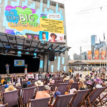 People at Federation Square in Melbourne seated in deck chairs in front of a small stage with a large billboard above. 