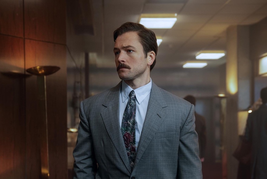 A man wearing a suit with short brown hair and a moustache walking forward in a dimly lit office corridor.