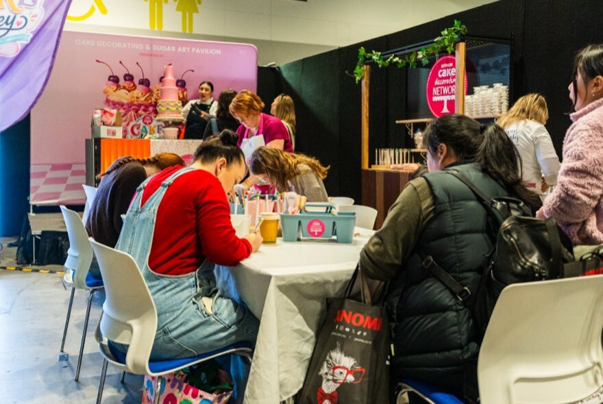 People seated at a long, white-clothed table, engaging in a hands-on activity inside an exhibition booth at a craft fair.