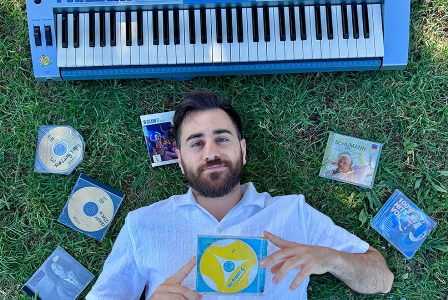 Person lying on the grass surrounded by CDs and an electric keyboard.