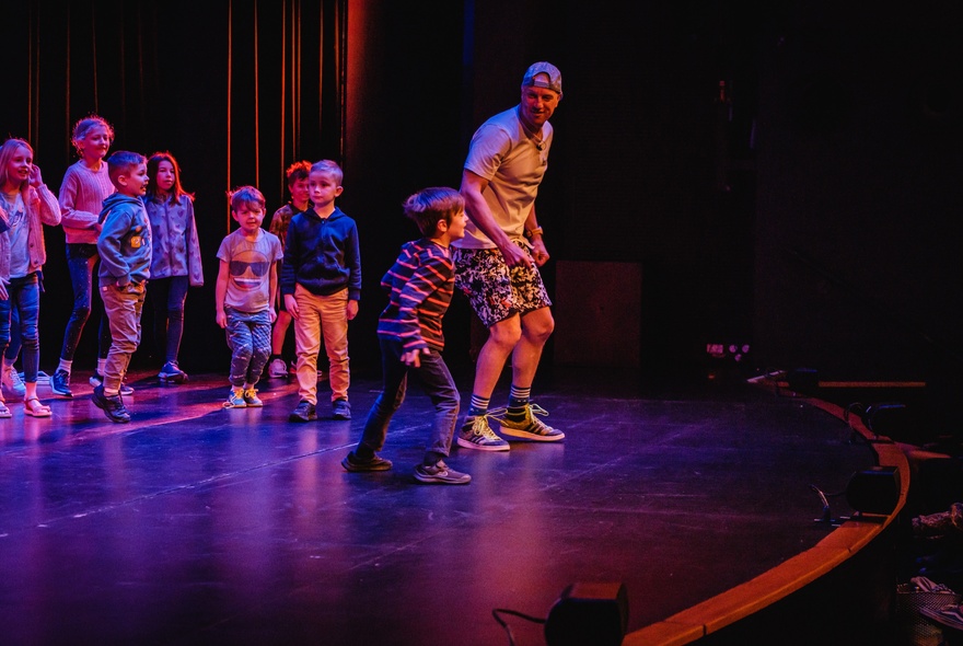 An adult and a group of primary school aged children on a theatre stage, interacting with each other.