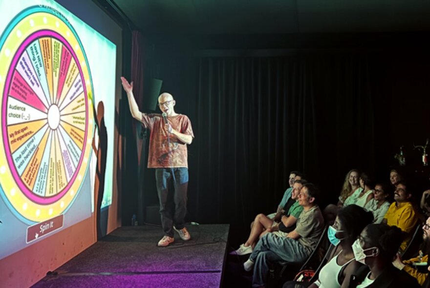 A comedian on stage in front of a projected spinning wheel with an audience looking on. 