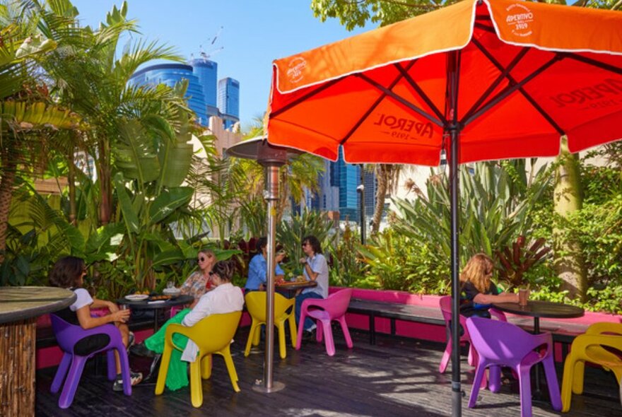 People sitting at colourful tables on a rooftop filled with palm trees.