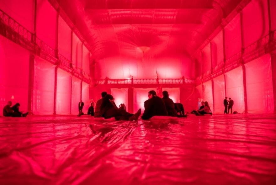 The red-lit interior of the Royal Exhibition Building, with people seated on the plastic-covered floor.