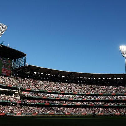 A wide-angle shot of the Melbourne Cricket Ground (MCG) grandstands filled with a large crowd under a clear, bright blue sky and two tall, stadium floodlight towers. 