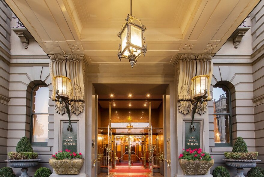 The grand entrance to Windsor Hotel, with open glass doors, hanging lights, sculptural architectural details and potted plants.