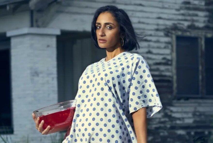 A woman with short dark hair in a spotted hospital gown, holding a bowl of red jelly.