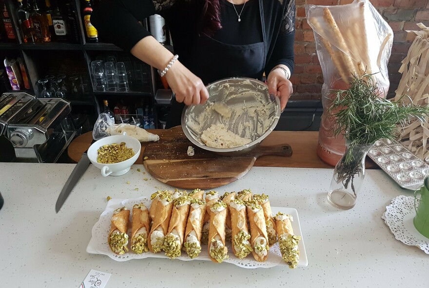 Hands holding a mixing bowl stirring cannoli mixture on a tabletop, with bowl of pistachios to the side.