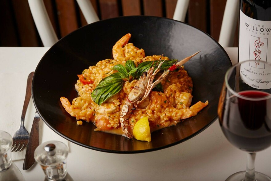 Table setting with wine bottle and glass, black dish with paella and cutlery.