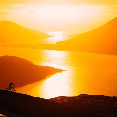 Mountains and a wide river at sunset with a lone cyclist on a path on the foreground.