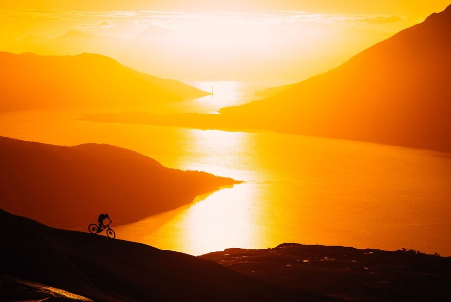 Mountains and a wide river at sunset with a lone cyclist on a path on the foreground.