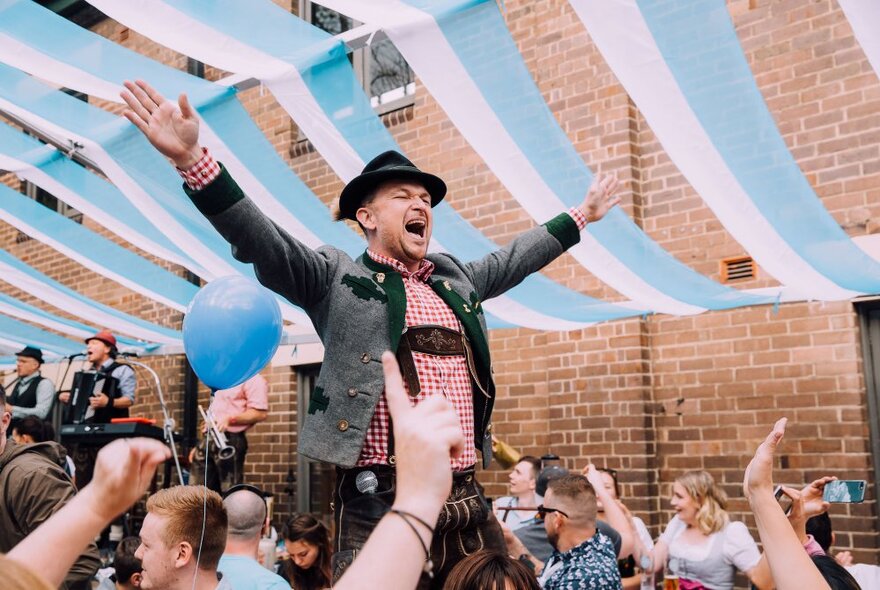 A man in a traditional German outfit and hat, standing on a table and yelling in a room full of people with streamers over the ceiling. 