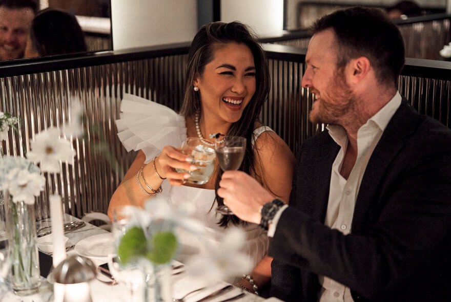 Two people clinking glasses in a toast while laughing at an elegant looking restaurant. 