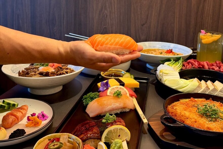 A table with multiple Japanese dishes and a hand holding a piece of salmon nigiri.