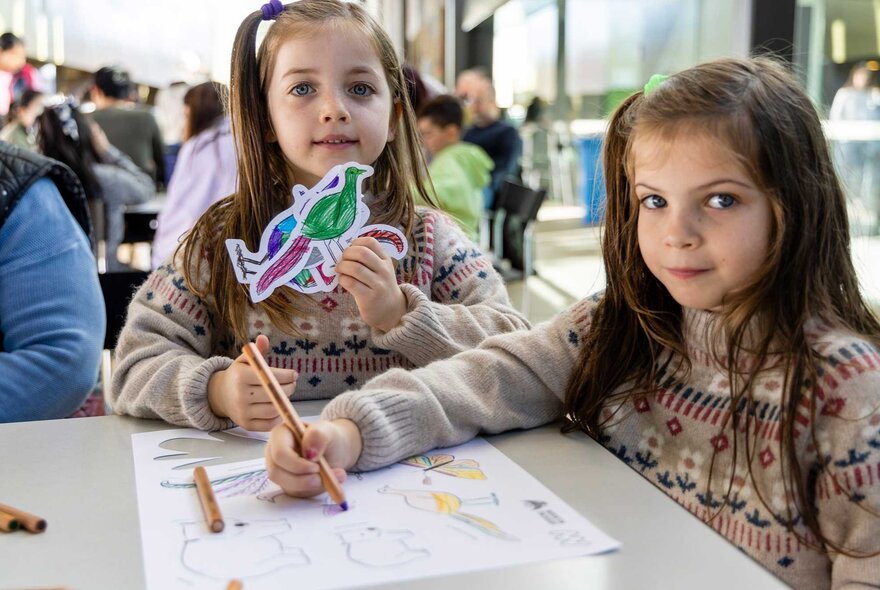 Two children seated at a table with pencils and paper, holding a bird activity cutout.