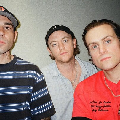 The three male members of the rock band DMA's in a room, all looking directly at the viewer, two of them wearing caps.