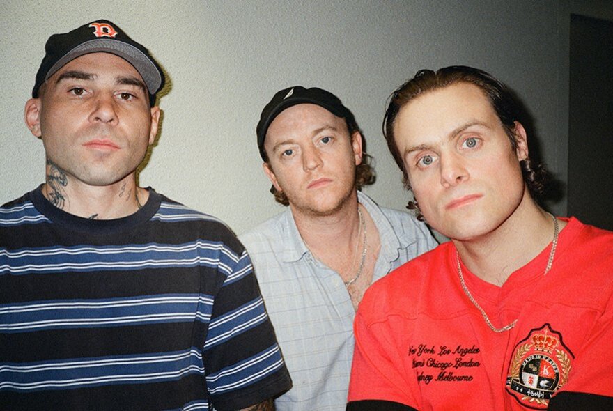 The three male members of the rock band DMA's in a room, all looking directly at the viewer, two of them wearing caps.